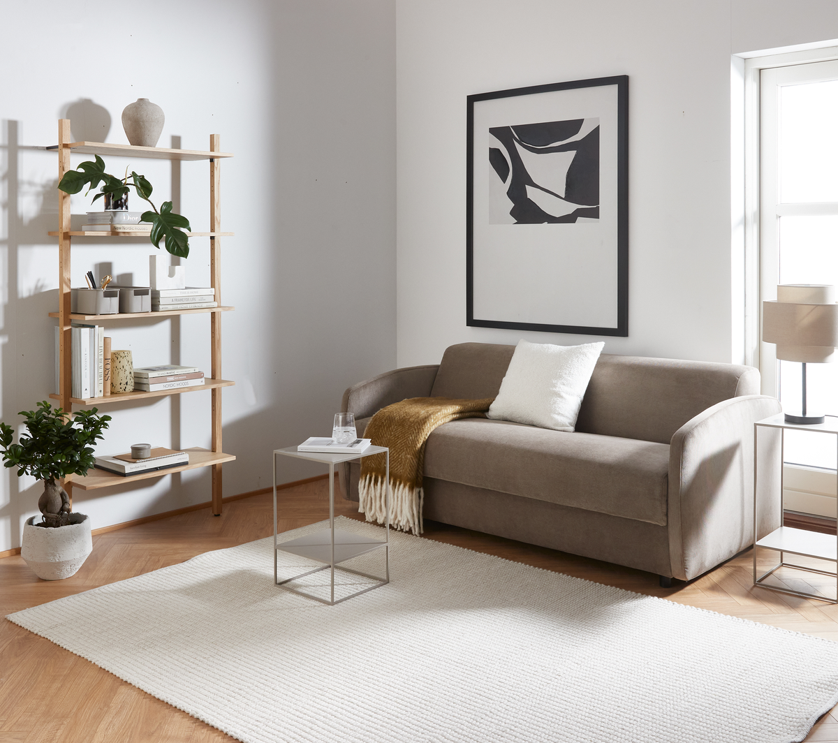 Lyst stue i skandinavisk stil med beige sofa, hvid pude og sennepsgul plaid, lille metalbord, reol med bøger og planter, stor lys tæppe og abstrakt vægbillede ved et vindue.