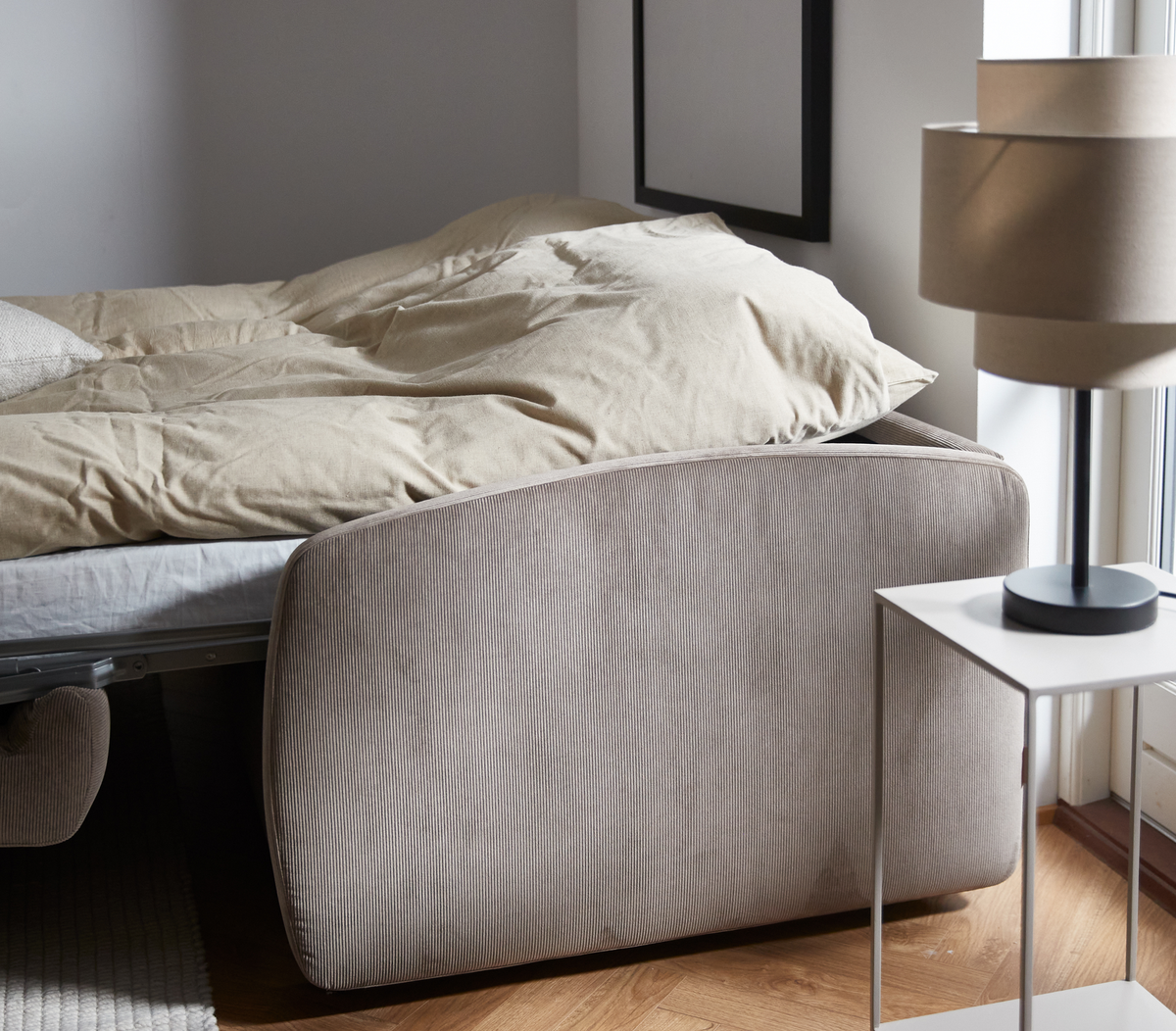 Seng med beige dyne ved siden af et hvidt sengebord med beige bordlampe ved et vindue; lyst trægulv og neutral indretning.