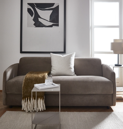 Beigefarvet sofa med hvid pude og brun plaid med frynser, lille sidebord med glas og bøger, abstrakt vægbillede og dagslys fra vindue.