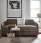 Beigefarvet sofa med hvid pude og brun plaid med frynser, lille sidebord med glas og bøger, abstrakt vægbillede og dagslys fra vindue.