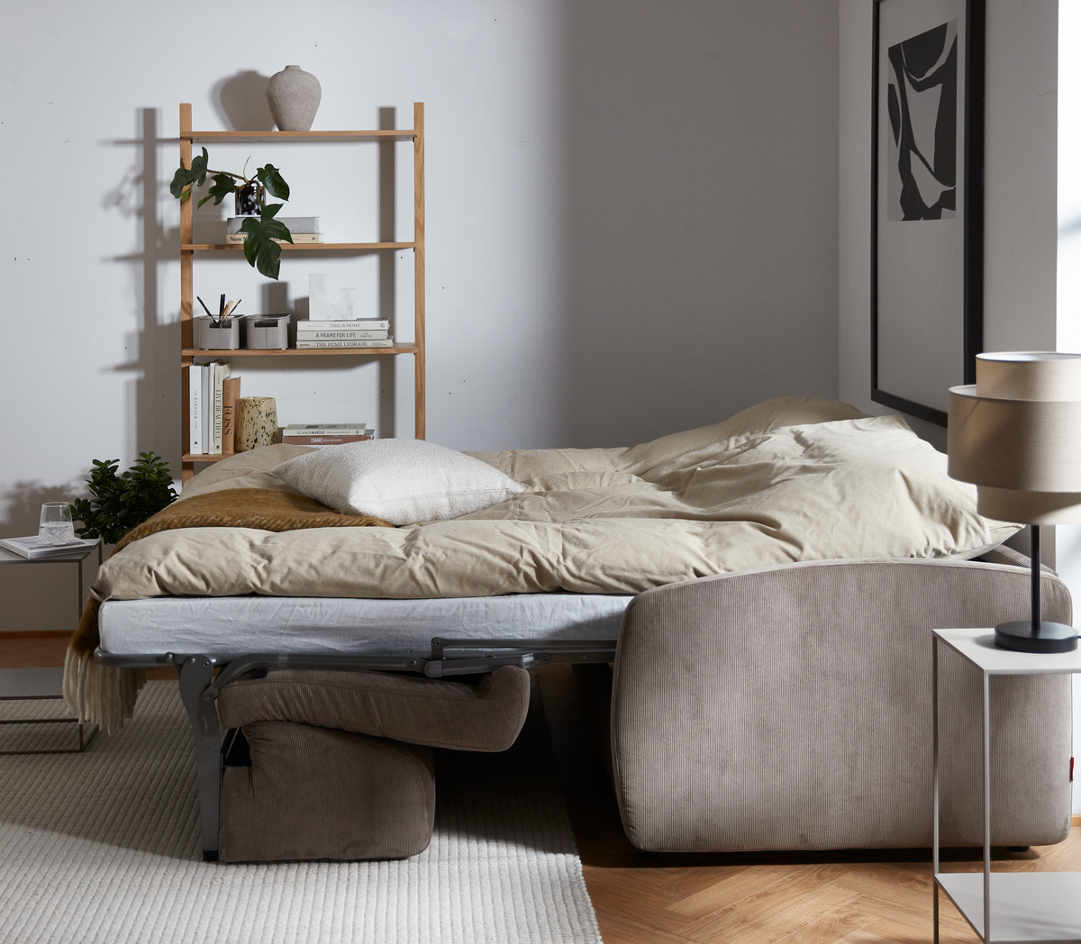 Udslået sovesofa i beige med sidebord og bordlampe, reol med bøger og planter i et lyst opholdsrum.