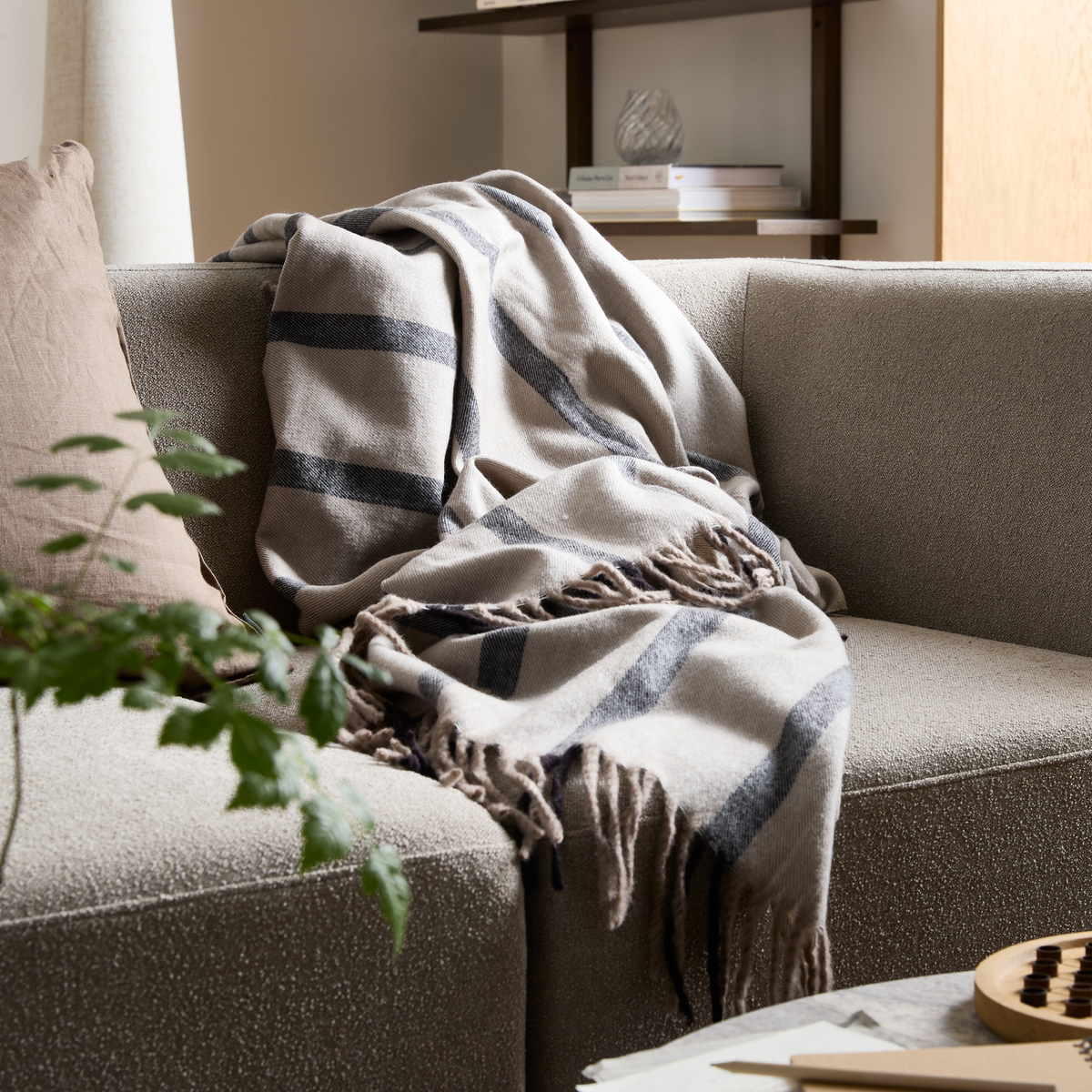 Beige sofa med blød, stribet plaid med frynser; pude, grøn plante og hylde i baggrunden i en rolig stue i dagslys.