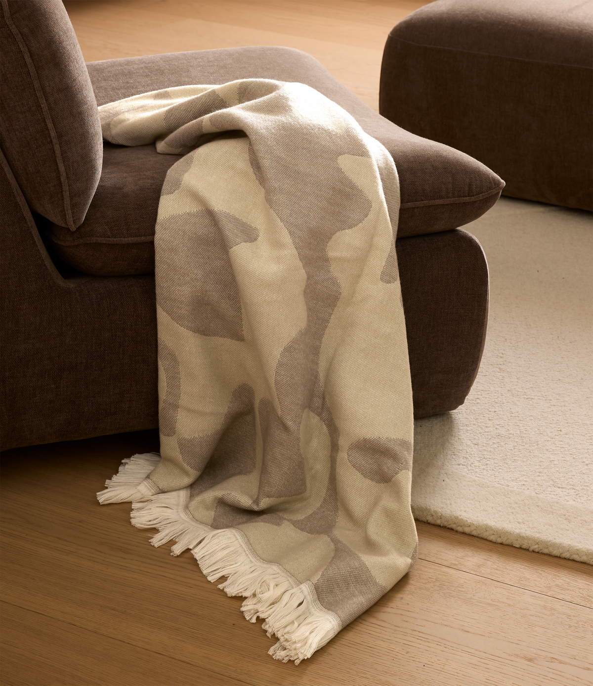 Beige og sandfarvet plaid med frynser over en brun sofa på trægulv ved siden af et lyst tæppe.