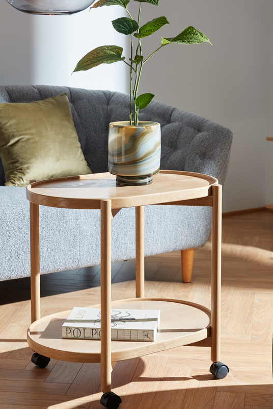 Lyst træ-sidebord på hjul med to hylder; en marmoreret glasvase med grøn plante står øverst, og bøger med titlen “POLPO” ligger på den nederste hylde, ved siden af en grå sofa i en solbeskinnet stue.