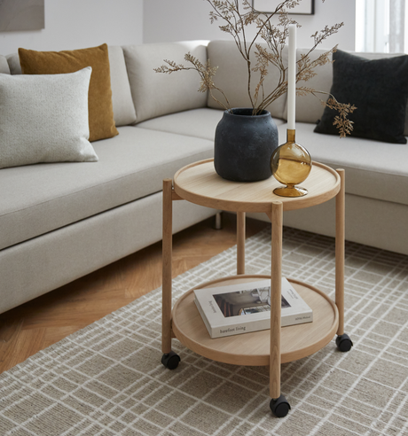 Lyst stue med beige sofa og rundt sidebord i eg på hjul; øverst en sort vase med tørrede grene og en ravfarvet lysestage, på nederste hylde en bog med titlen ”barefoot living”.