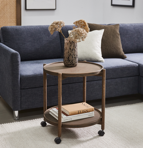 Rundt sidebord i træ på hjul med en spættet vase med tørrede blomster og bøger som FOSS og barefoot living på den nederste hylde ved siden af en blå sofa med puder på et lyst tæppe.
