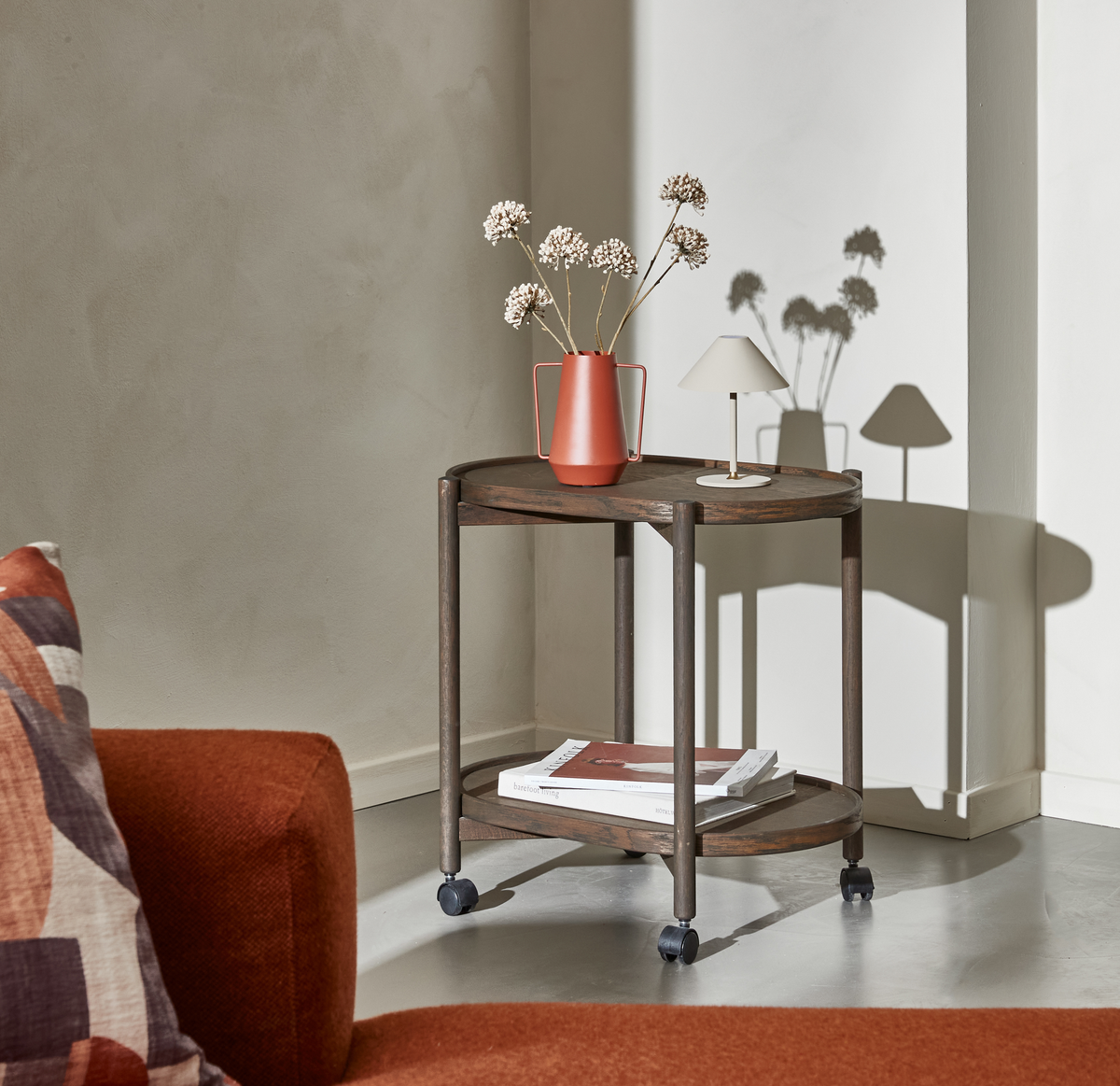Solbelyst hjørne med lille sidebord/rullevogn i mørkt træ, vase med tørrede blomster og lille hvid bordlampe; bøger på nederste hylde og en rustfarvet sofa i forgrunden.