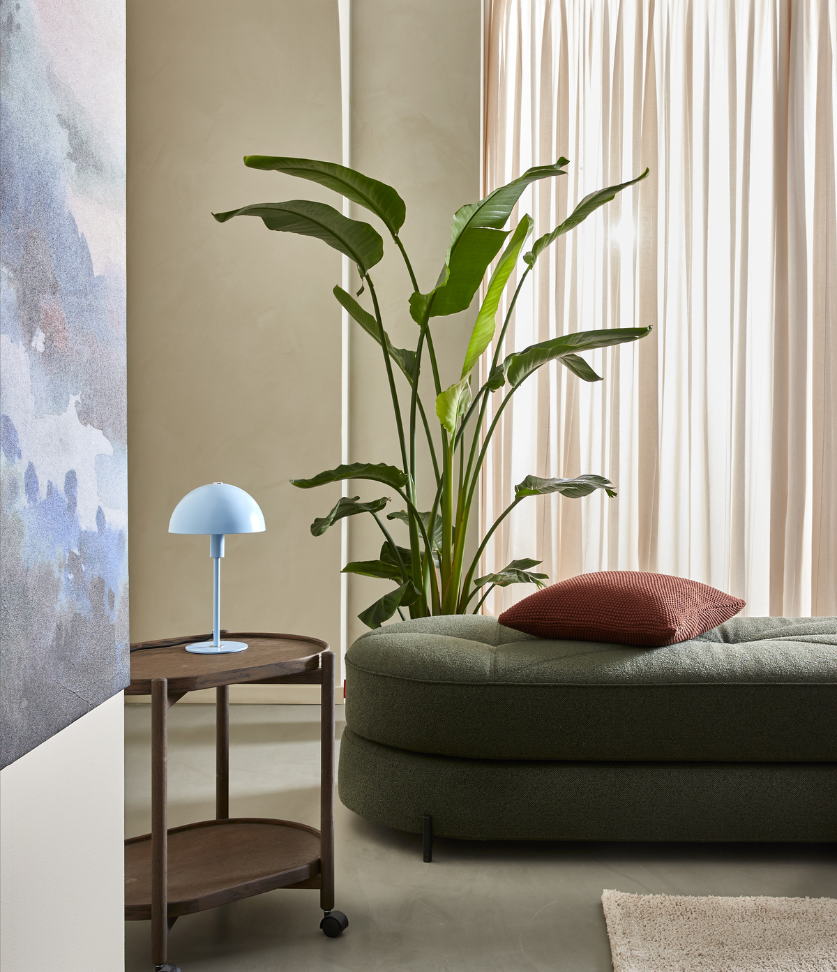 Lyst stue med grøn daybed, rød pude, stor potteplante, beige gardiner og blå bordlampe på træserveringsvogn.