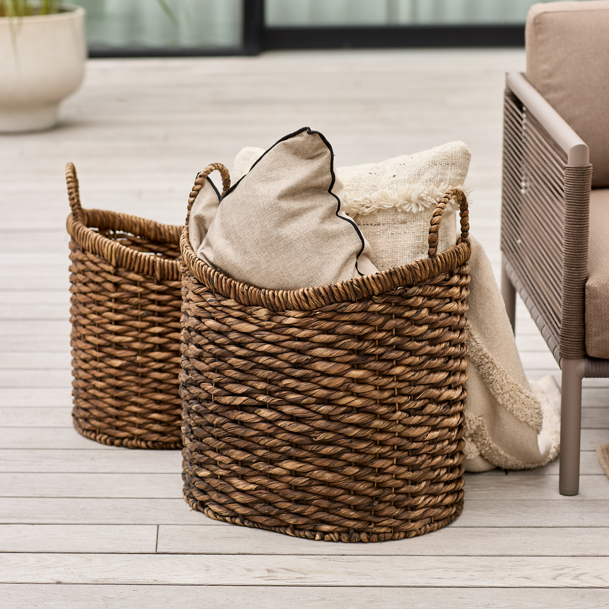 To flettede kurve på en terrasse, fyldt med beige puder og et cremefarvet tæppe ved siden af en loungestol.