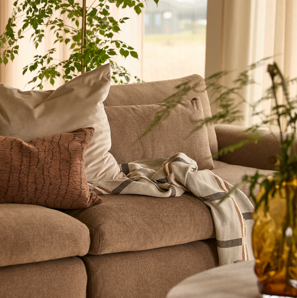 Nærbillede af en beige sofa med neutrale puder, et stribet plaid over armlænet, grønne planter og en ravfarvet vase i blødt dagslys.