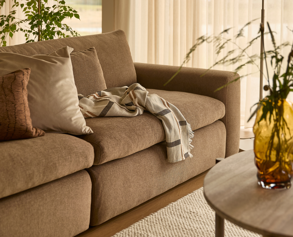 Stue med brun sofa, puder og plaid, gul glasvase med grønne kviste på sofabord, lyse gardiner og tekstureret tæppe i blødt dagslys.