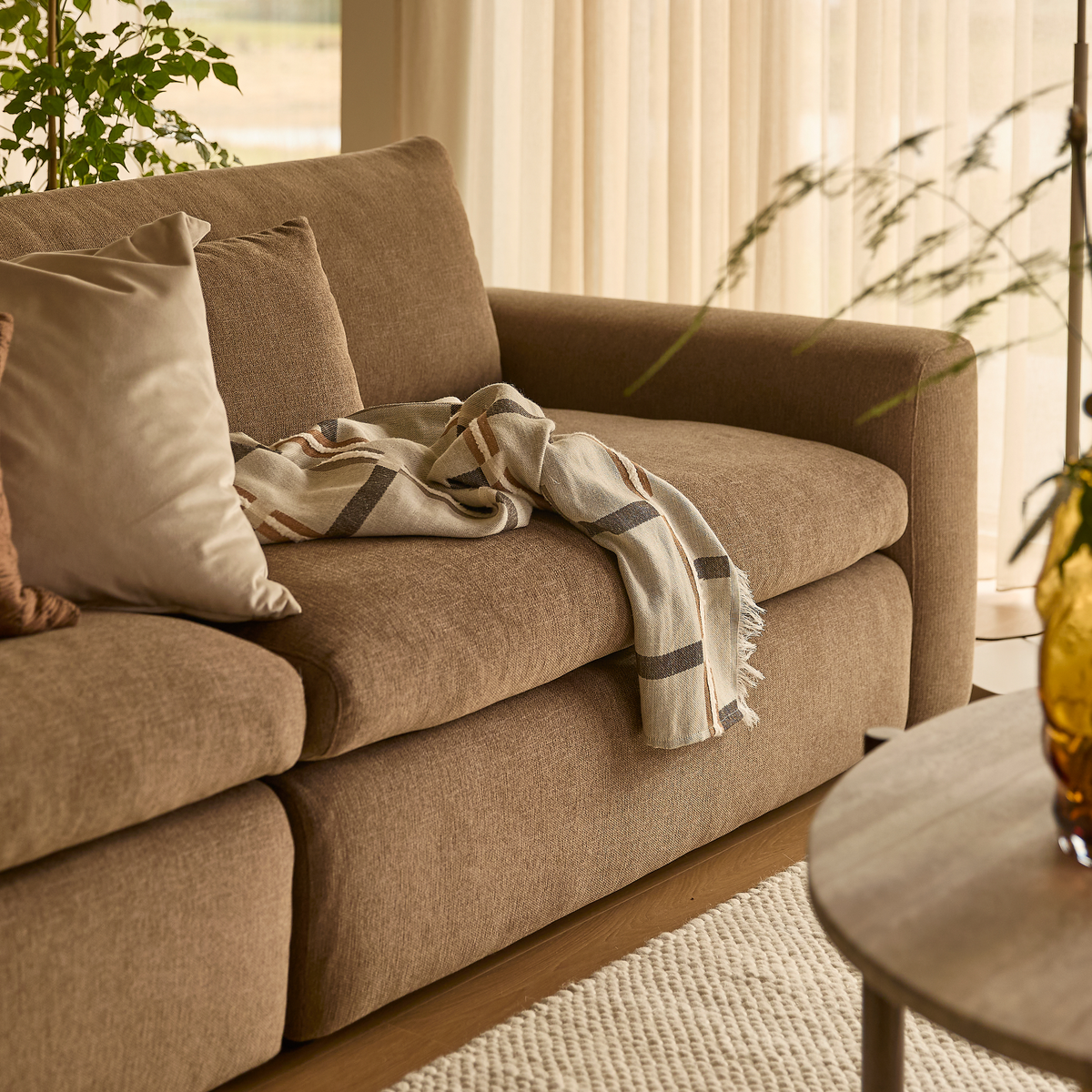 Brun sofa med beige pude og mønstret plaid, lyse gardiner, grønne planter og ravfarvet vase på sofabord i en varm, skandinavisk stue.