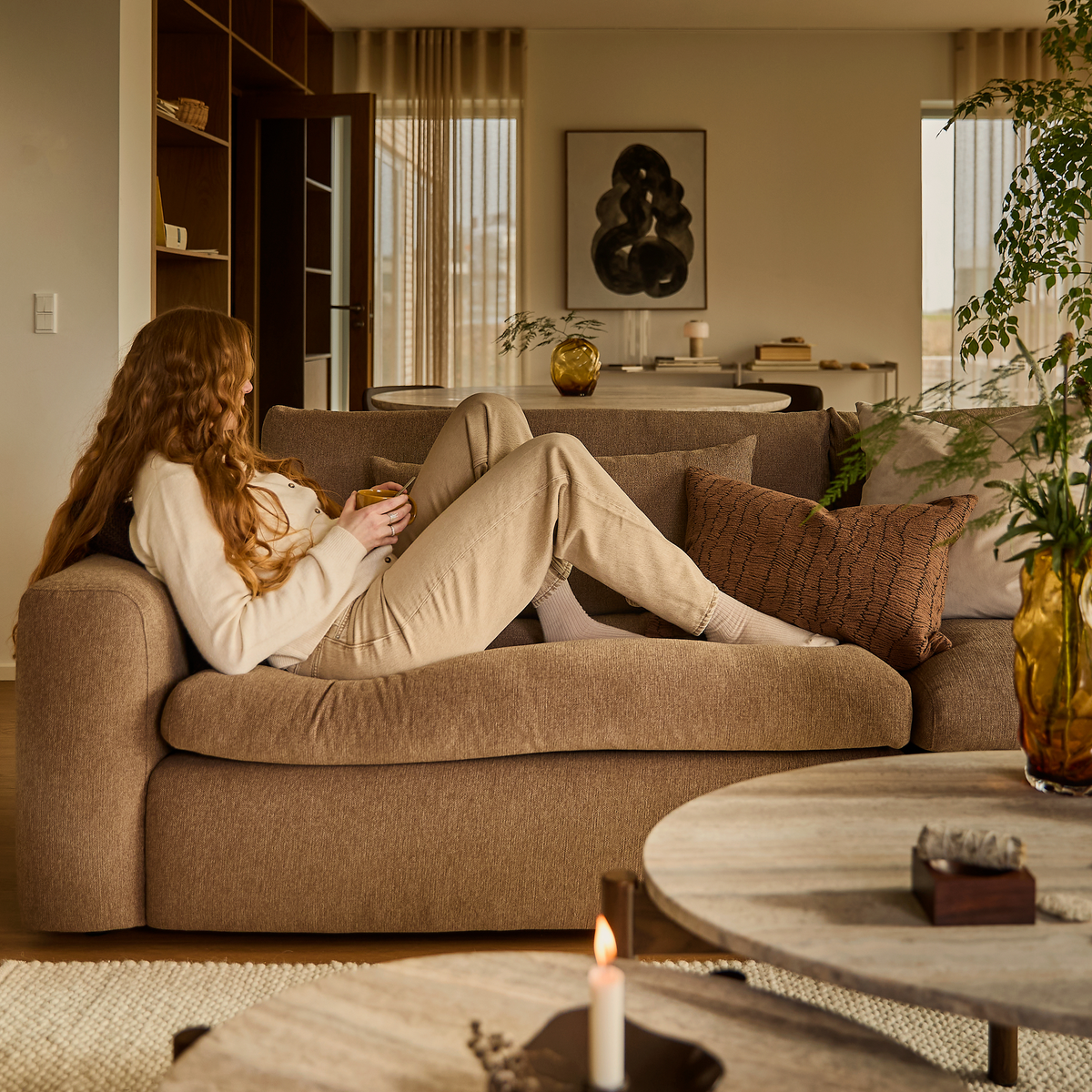 Person ligger afslappet på en beige sofa i et lyst stue-miljø med puder, planter, stearinlys og kunst på væggen.