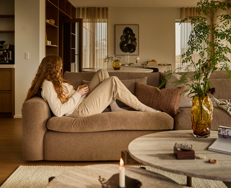 Person ligger afslappet på en beige sofa i en varm, skandinavisk stue med grønne kviste i ravfarvet vase, tændt lys og runde sofaborde.