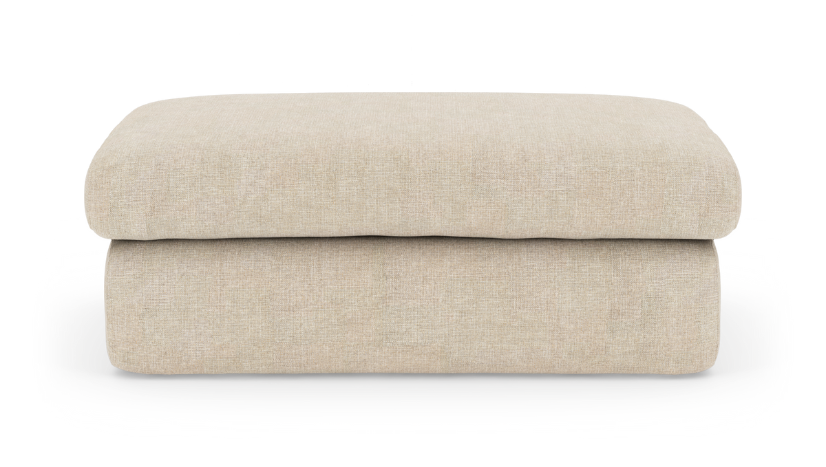 Beige polstret puf/skammel med blød, lav profil på hvid baggrund set forfra.