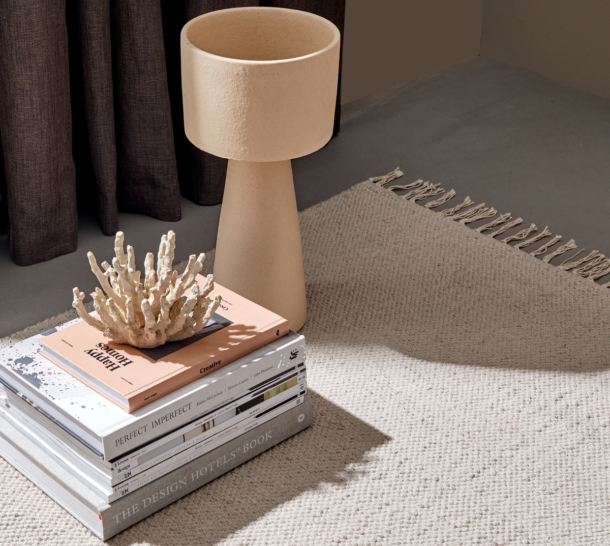 En stak designbøger, bl.a. “THE DESIGN HOTELS BOOK”, “PERFECT IMPERFECT” og “Creative”, toppet af en koralskulptur ved siden af en beige tekstureret bordlampe på et neutralt vævet tæppe med frynser; blødt dagslys og mørke gardiner i baggrunden.
