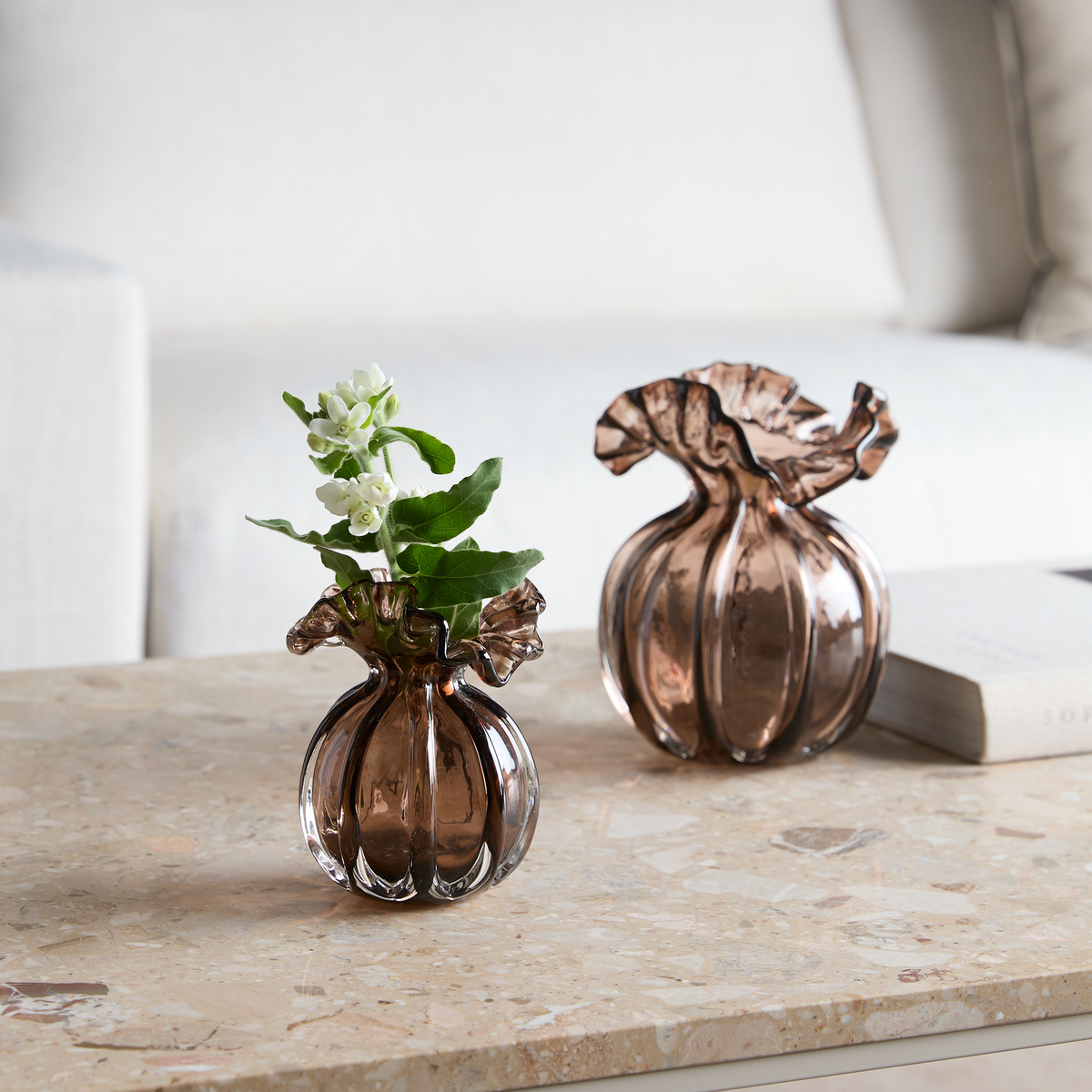 To små riflede glasvaser i røgbrun med bølget åbning på en beige stenbordplade; den ene med hvide blomster og grønne blade; sofa og bog i baggrunden.
