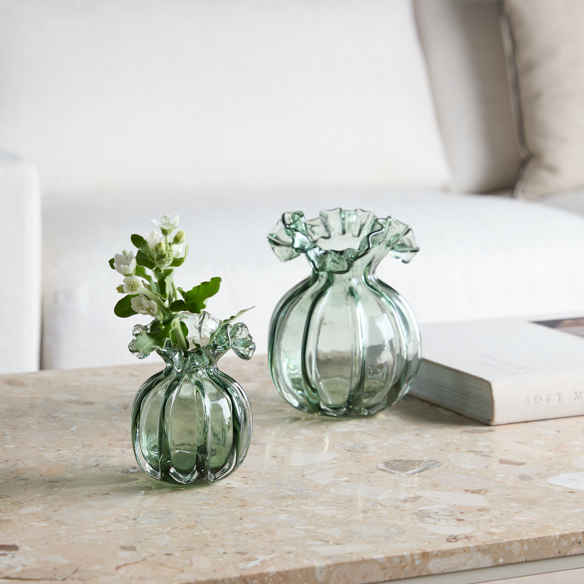 To grønne, riflede glasvaser med bølget kant står på et stenbord; den lille har hvide blomster, en bog ligger ved siden af og en sofa ses i baggrunden.