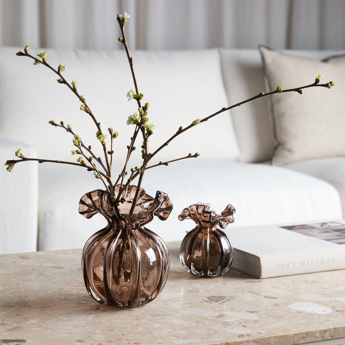 Brun røgfarvet glasvase med bølget kant og blomstrende grene på et sten-sofabord, ved siden af en mindre vase og en bog, Soft Minimal, med lys sofa i baggrunden.