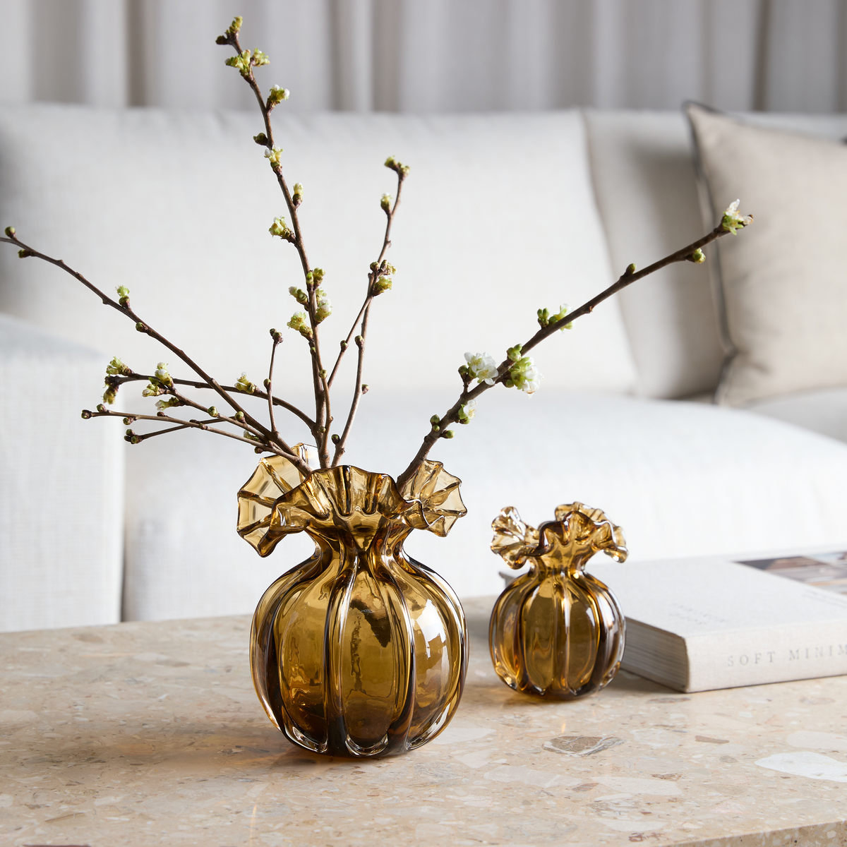 Ravfarvede glasvaser med ruflet kant på et stenbord; den store rummer grene med hvide knopper, ved siden af en bog med titlen Soft Minimal og en lys sofa i baggrunden.