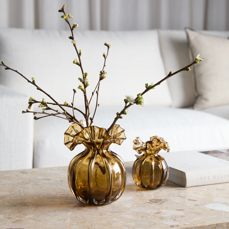 Ravfarvede glasvaser med ruflet kant på et stenbord; den store rummer grene med hvide knopper, ved siden af en bog med titlen Soft Minimal og en lys sofa i baggrunden.