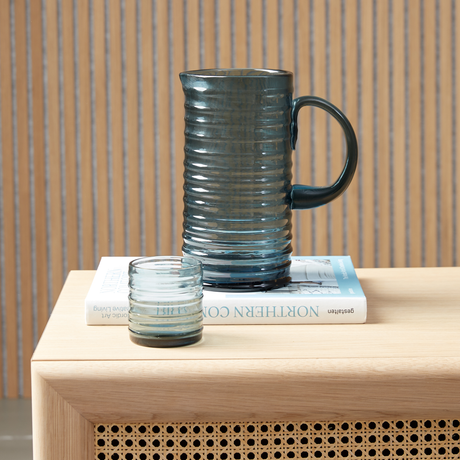 Riflet blå glaskande med matchende lille tumbler på en bog med titlen Northern Comfort, gestalten, på en lys træoverflade foran en lamelvæg.