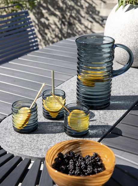 Blå riflet glaskande med citronskiver og tre matchende glas på et gråt udendørsbord; skål med brombær i forgrunden.