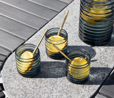 Tre riflede, røgfarvede glas med citronskiver og guldfarvede sugerør på en grå stenplade udendørs i sollys; matchende karaffel delvist i billedet.