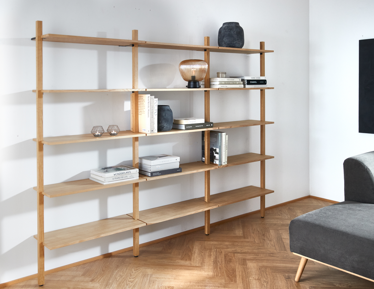 Lys egetræshylde mod hvid væg med bøger (bl.a. CHANEL, WONDERPLANTS), en sort vase og en lille bordlampe; grå sofa i hjørnet.