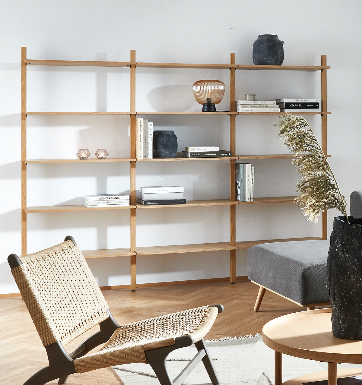 Lyst skandinavisk stue med åben reol i egetræ, bøger med titlerne CHANEL, NORDIC MOODS og WONDERPLANTS/A FRAME FOR LIFE, en bordlampe, sorte vaser og pampasgræs; grå sofa, fletstol og rundt sofabord.