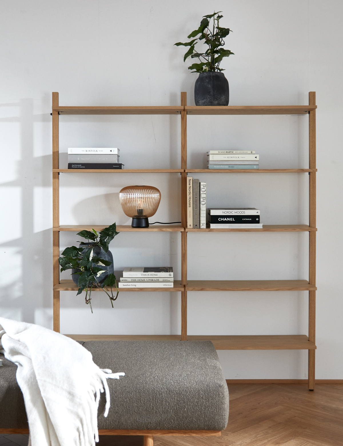 Lys skandinavisk indretning med åben træreol; planter, en riflede glasbordslampe og bøger som The Kinfolk, Scandinavian, Nordic Moods, CHANEL, This Is Home og The Home Upgrade; grå bænk med hvidt plaid foran.