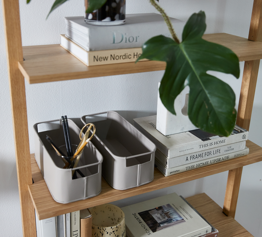 Reol i træ med beige opbevaringskasser med saks og penne, grønt planteblad og bogstakke med titlerne Dior, New Nordic Home, This Is Home, A Frame for Life, The Home Upgrade og Scandi Rustic.