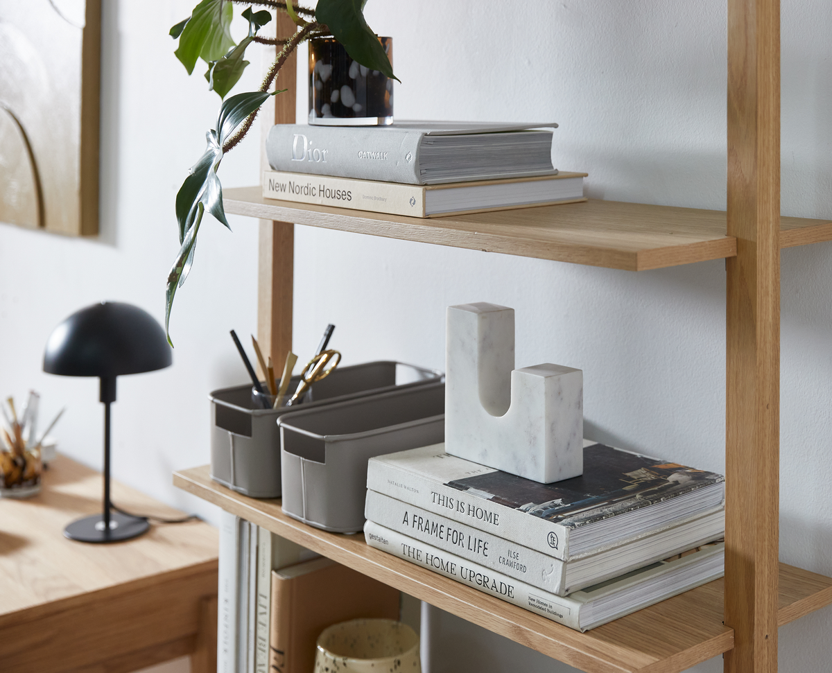 Nærbillede af reol i lys eg med designbøger – Dior, New Nordic Houses, This Is Home, A Frame for Life og The Home Upgrade – et U‑formet bogstøtte i marmor, grøn plante og sort bordlampe i et roligt hjemmekontor.