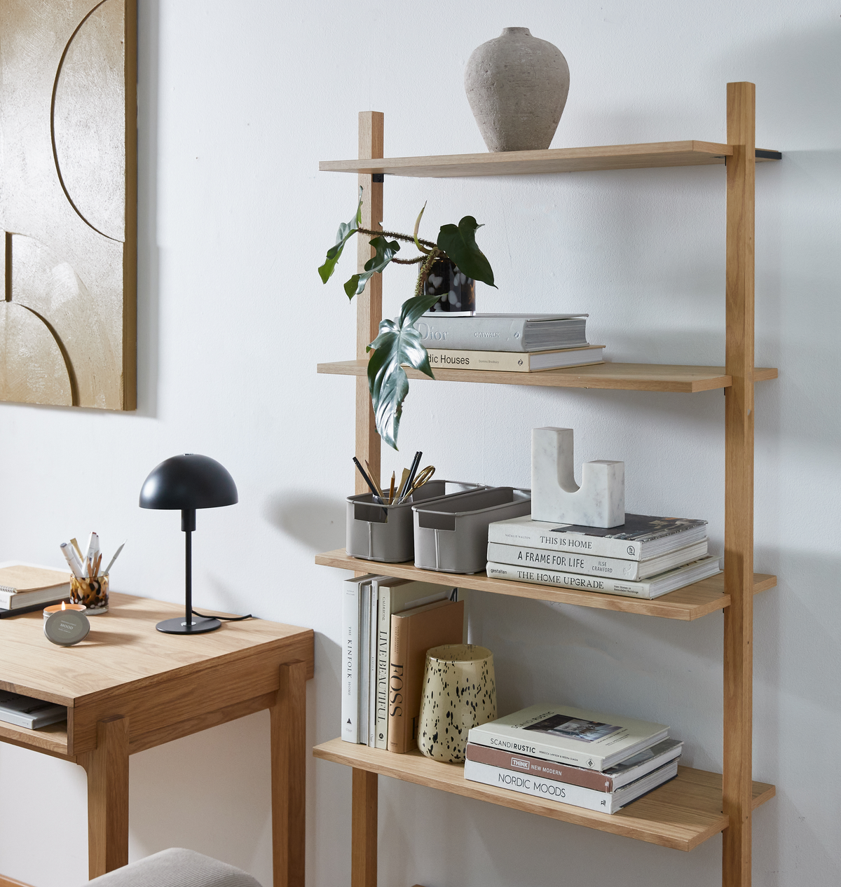 Lyst hjemmekontor med åbne træhylder med designbøger som “This Is Home”, “A Frame for Life”, “The Home Upgrade”, “Scandi Rustic” og “Nordic Moods”, en lille sort bordlampe, grå vase og grøn plante ved et skrivebord i lyse neutrale nuancer.