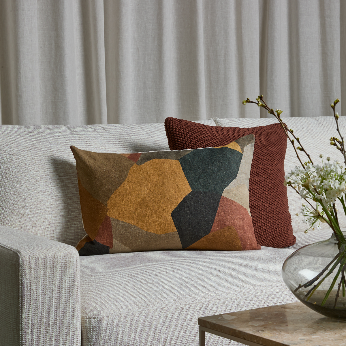 Lys sofa med to pyntepuder – en i rust og en med grafisk mønster i jordtoner – foran lyse gardiner; glasvase med blomstrende grene på sofabordet.