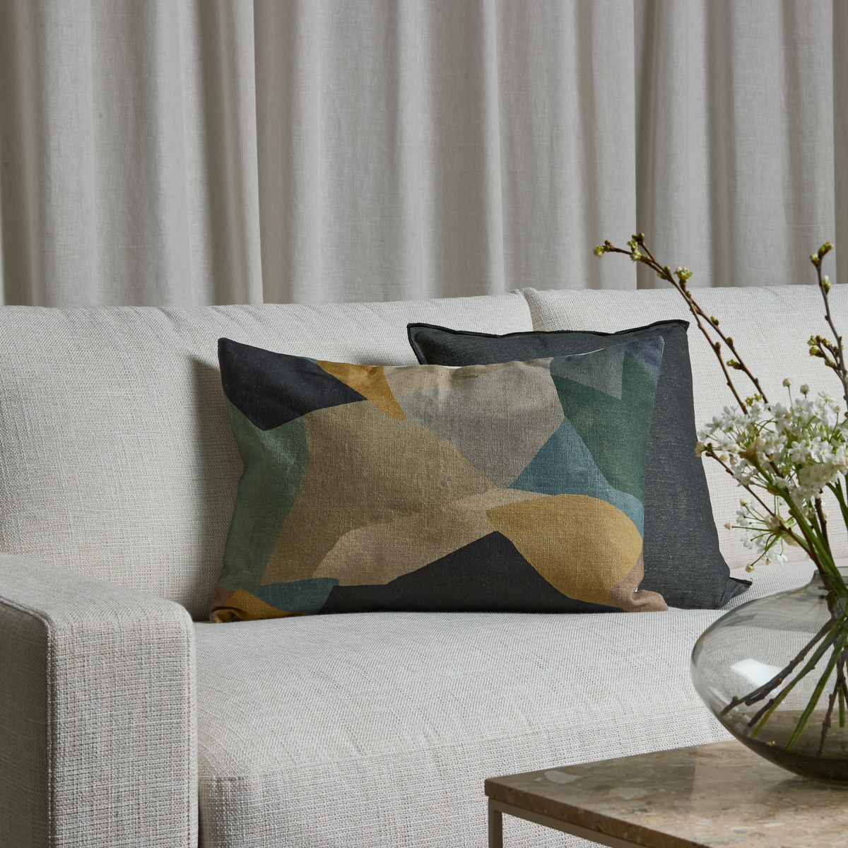 Lys beige sofa med to rektangulære pyntepuder med abstrakt mønster i mørkegrå, grøn og gul; foran står et sofabord med glasvase og hvide blomstergrene, med lyse gardiner i baggrunden.
