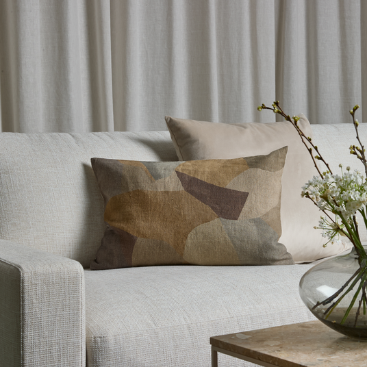 Lys sofa med to puder, en med abstrakt mønster i jordtoner, foran lyse gardiner; glasvase med hvide grene på bordet.