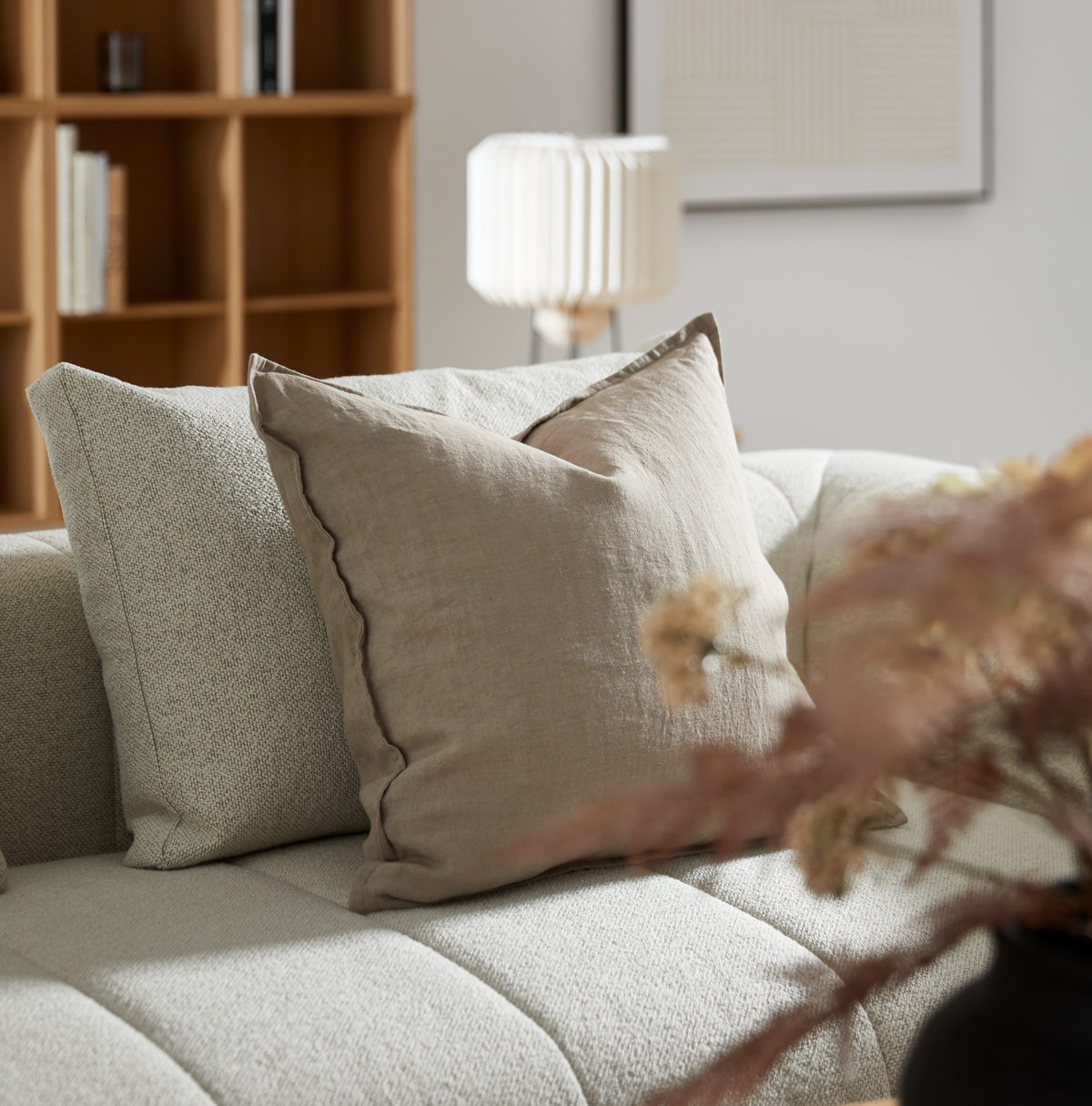 Nærbillede af en beige linnedpude og en lysgrå pude på en lys sofa i stuen; træreol, hvid plisseret bordlampe og tørrede blomster i forgrunden.