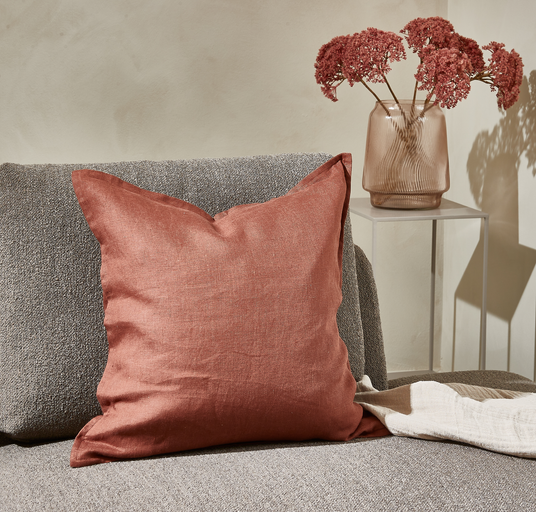 Rustfarvet hørpude på grå sofa ved siden af glasvase med tørrede blomster.