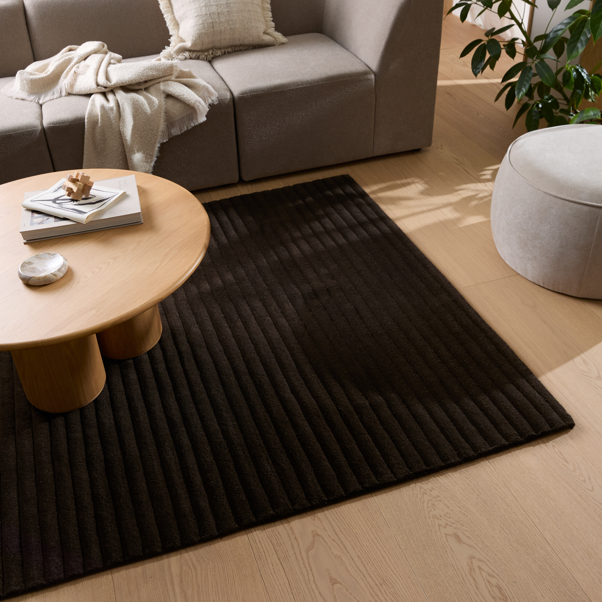 Beige sofa med plaid og pude, rundt sofabord i eg med bøger, mørkt rillet tæppe på lyst trægulv, puff og grøn plante i en moderne stue.