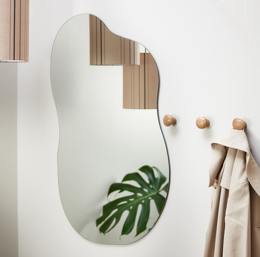 Organisk vægspejl på hvid væg med runde træknager, beigefarvet frakke og en monstera i spejlet; pendellampe ses i refleksionen.