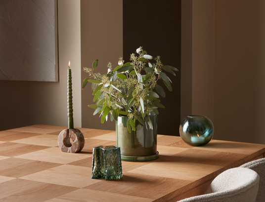Stilleben på træbord med grøn vase med eukalyptus, tændt grønt spiralstearinlys i marmorlysestage og to grønne glaslysholdere mod beige væg.