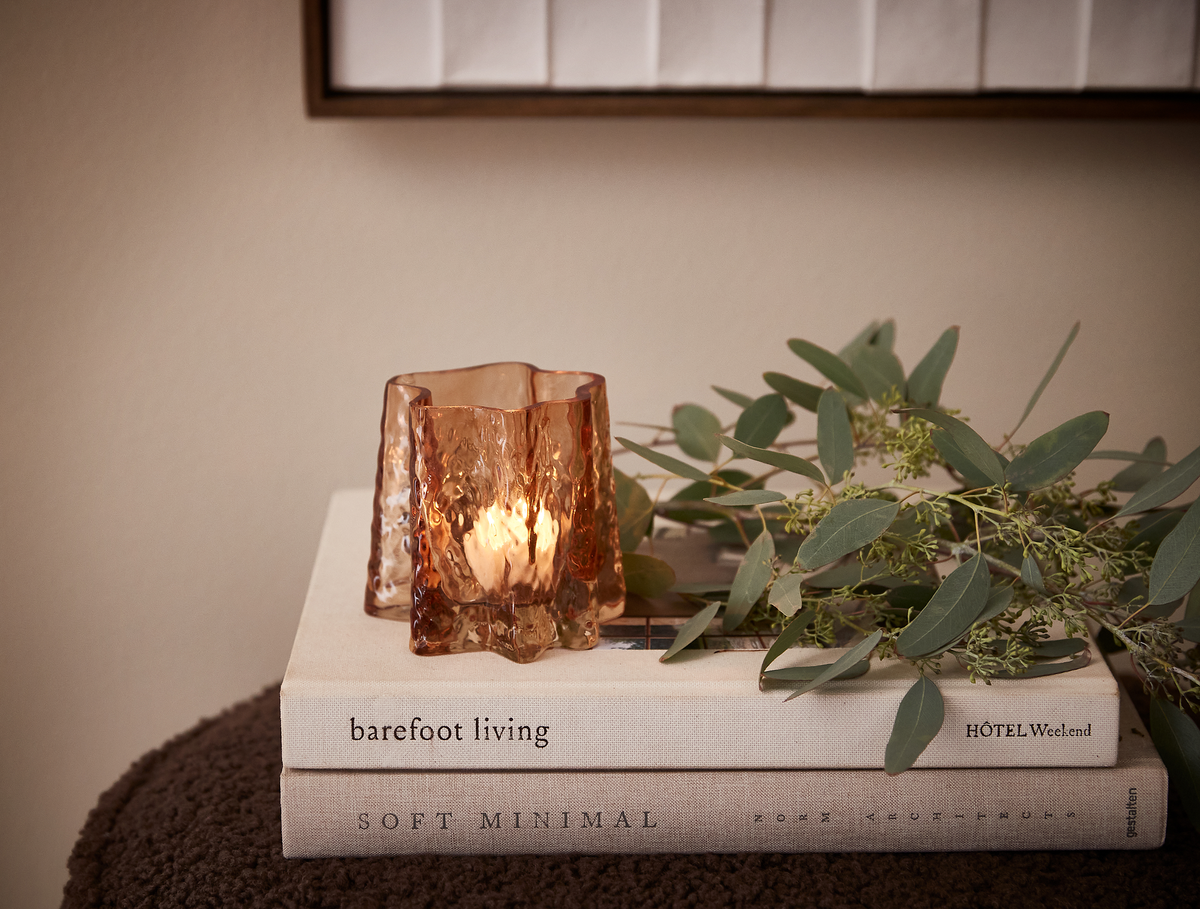 Ravfarvet glaslysestage med varm glød på to indretningsbøger – barefoot living og SOFT MINIMAL – sammen med en eukalyptusgren; HÔTEL Weekend i hjørnet.