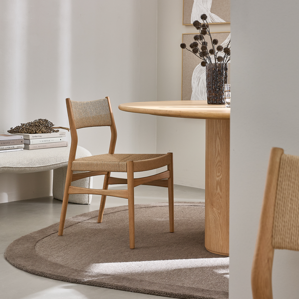 Lyst spisekrog i neutrale toner med rundt egetræsbord, vævet træstol på beige tæppe, vase med tørrede kviste, vægkunst og en stak Kinfolk-bøger.