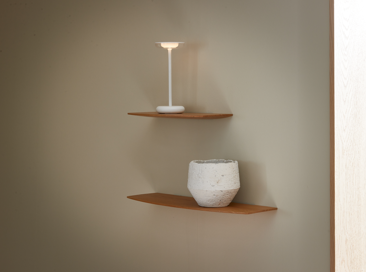 To væghylder i træ med en hvid bærbar bordlampe på den øverste og en hvid struktureret vase på den nederste, mod en beige væg.