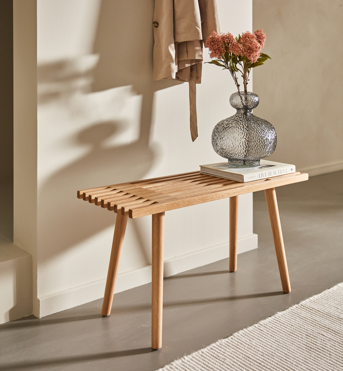 Lys træbænk mod beige væg med struktureret glasvase med lyserøde blomster på en stak bøger; blødt dagslys og en hængt frakke kaster skygger.