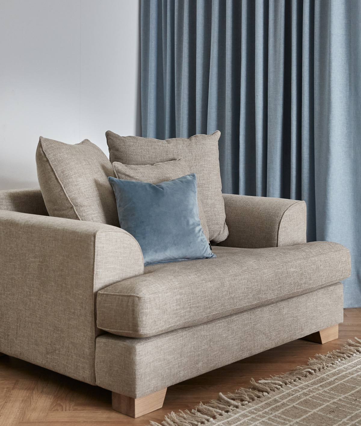 Beige stofsofa med blå velourpude foran gråblå gardiner og et frynset tæppe på trægulv.