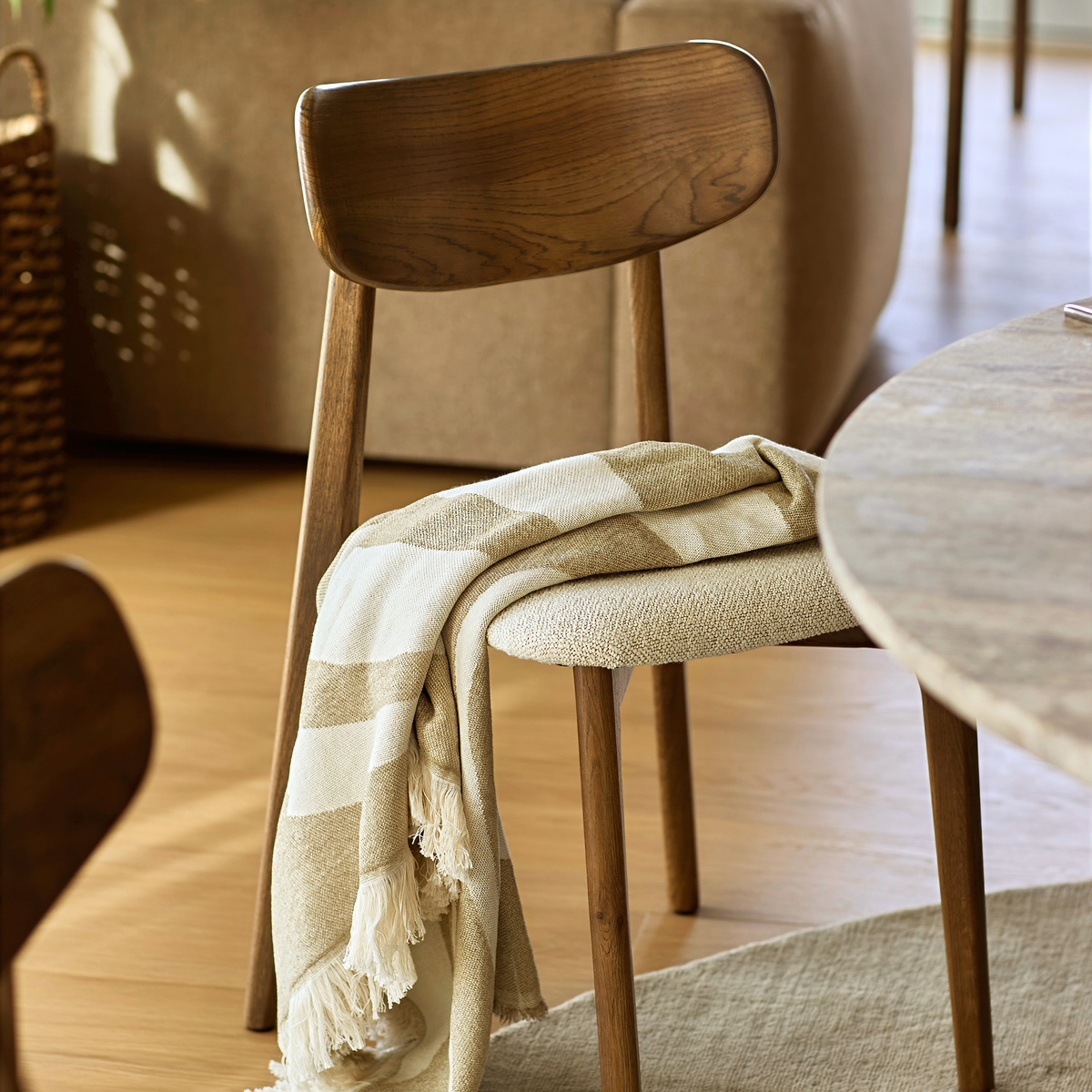 Træstol ved rund bordplade med beige‑hvid stribet plaid med frynser lagt over sædet i et lyst, varmt spiseområde.