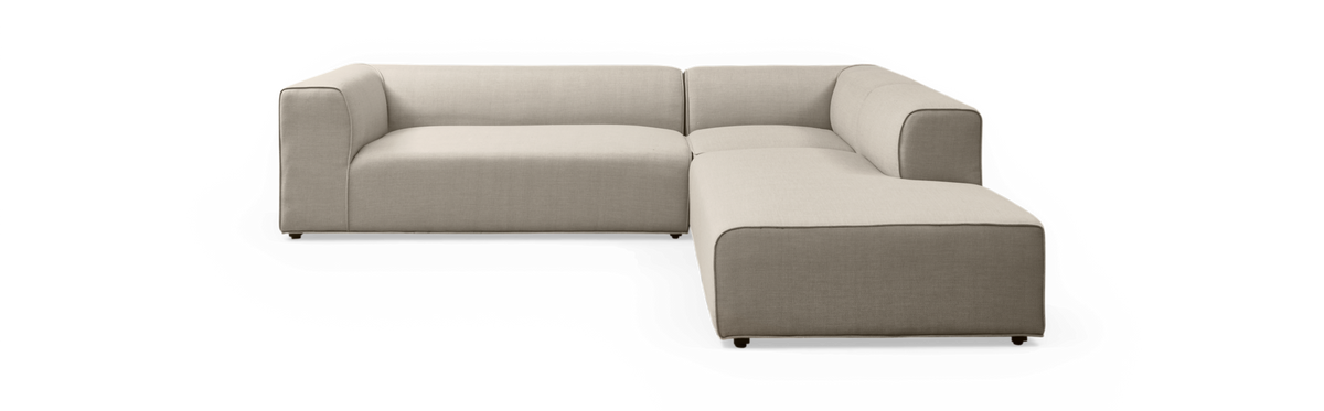 L-sofa i beige stof med chaiselong til højre, frilagt mod sort baggrund.