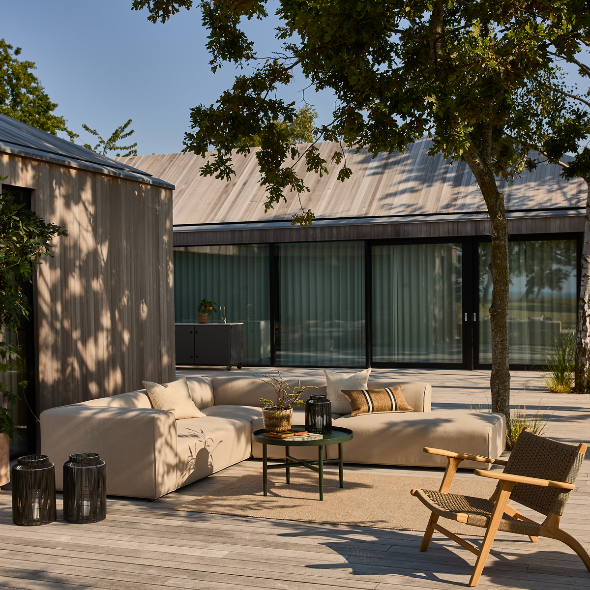 Lys terrasse med beige modulsofa, runde sofaborde og træskygger foran et moderne hus med store glas-skydedøre.